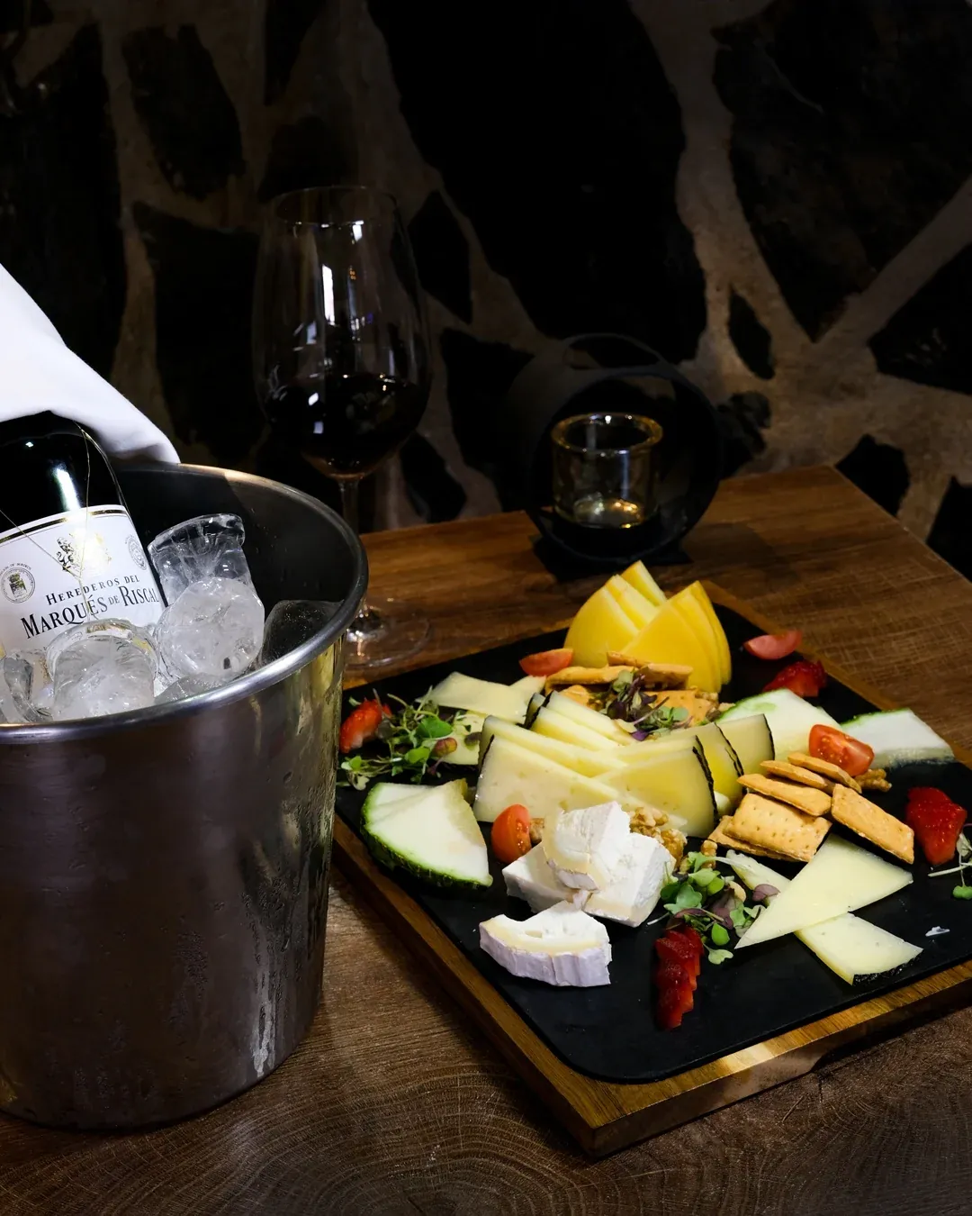 Tabla de quesos y una botella de vino en cubitera, con la pared de piedra del comedor al fondo